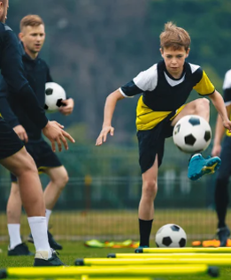 Junge in gelb-schwarzem Trikot trainiert Fußball mit mehreren Bällen und Hütchen auf einem Rasenplatz